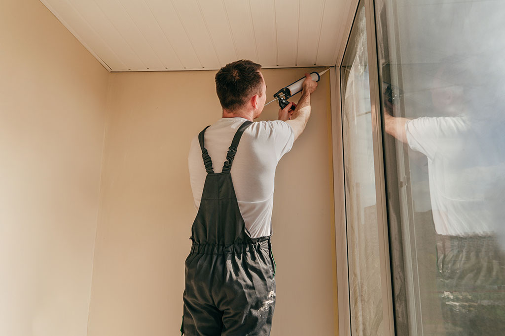 Young man wearing overalls sealing cracks between window and trim using waterproof silicone caulk on the balcony.